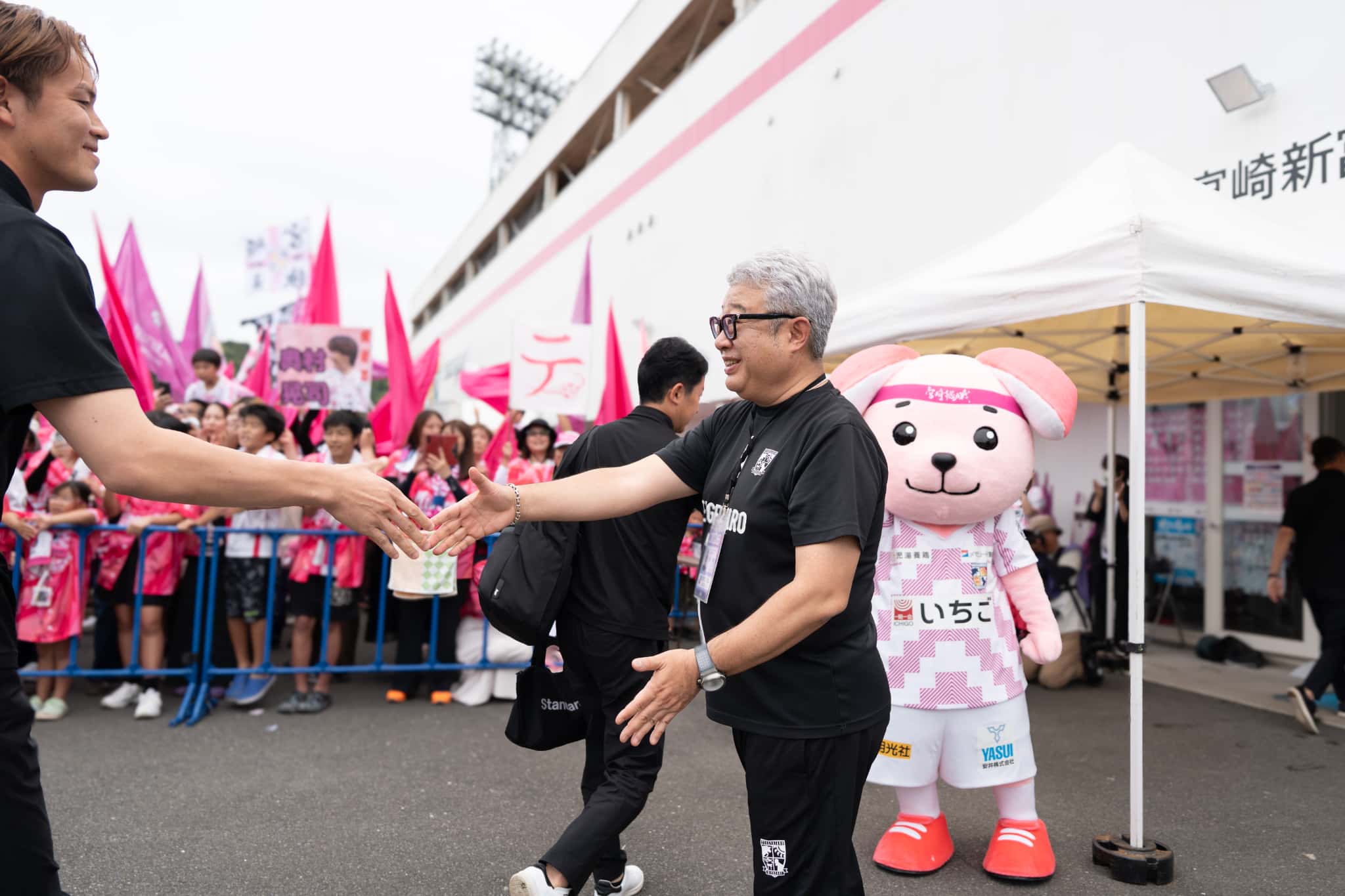 スタジアム入口前で到着した選手を出迎え握手する石原実。後方にサポーターとクラブマスコット