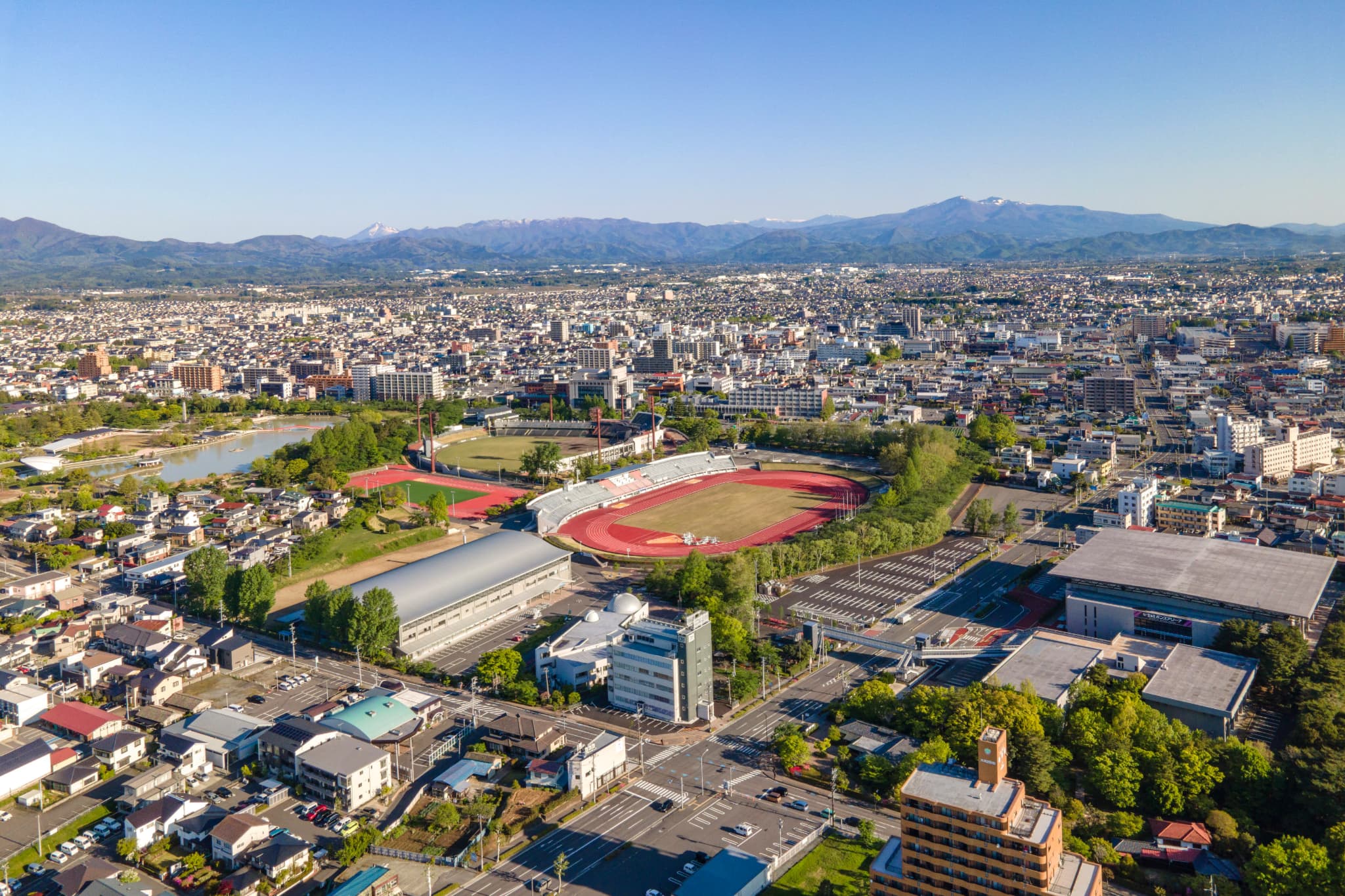 郡山市開成山地区の空撮。左手に池、手前に住宅地、中央に陸上競技場と体育館、奥に市街地と山並みが広がる