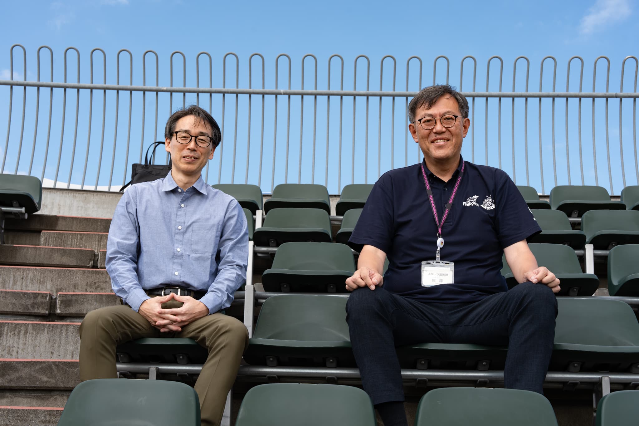 スタンドの座席に並んで座る郡山市選ばれるまち推進課の髙橋勇介（左）とスポーツ振興課の佐藤聡一（右）