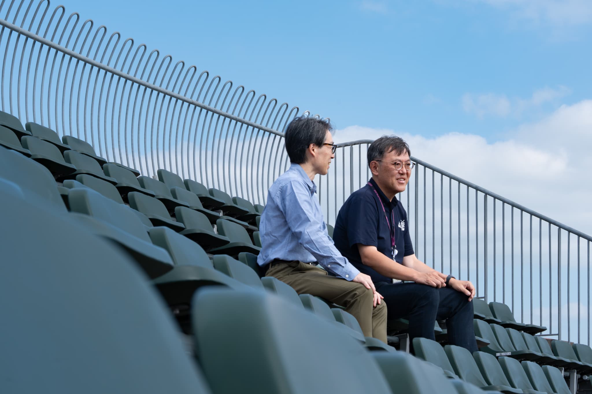 野球場のスタンドに並んで座る郡山市選ばれるまち推進課の髙橋勇介とスポーツ振興課の佐藤聡一