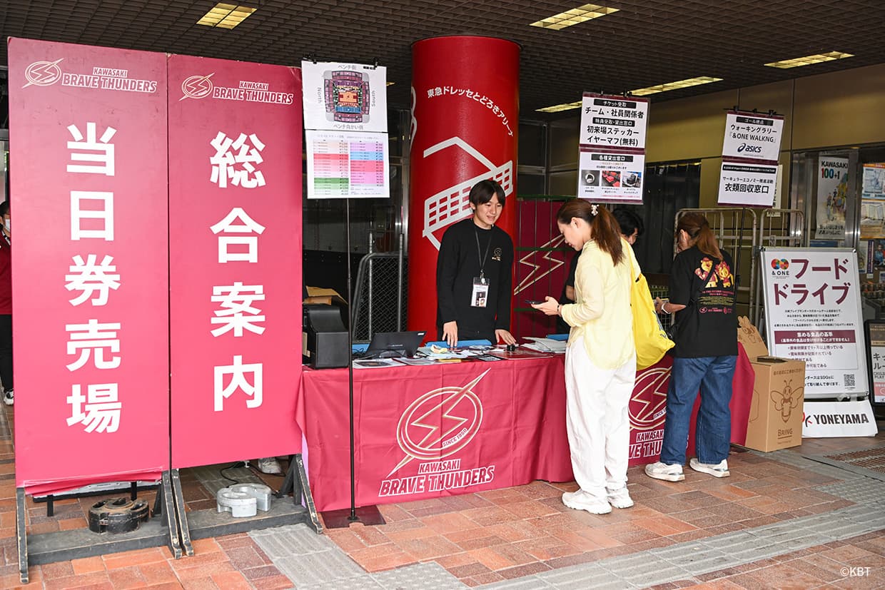 「当日券売場」「総合案内」ののぼりの横で、川崎ブレイブサンダースのテーブルに立つスタッフと来場者