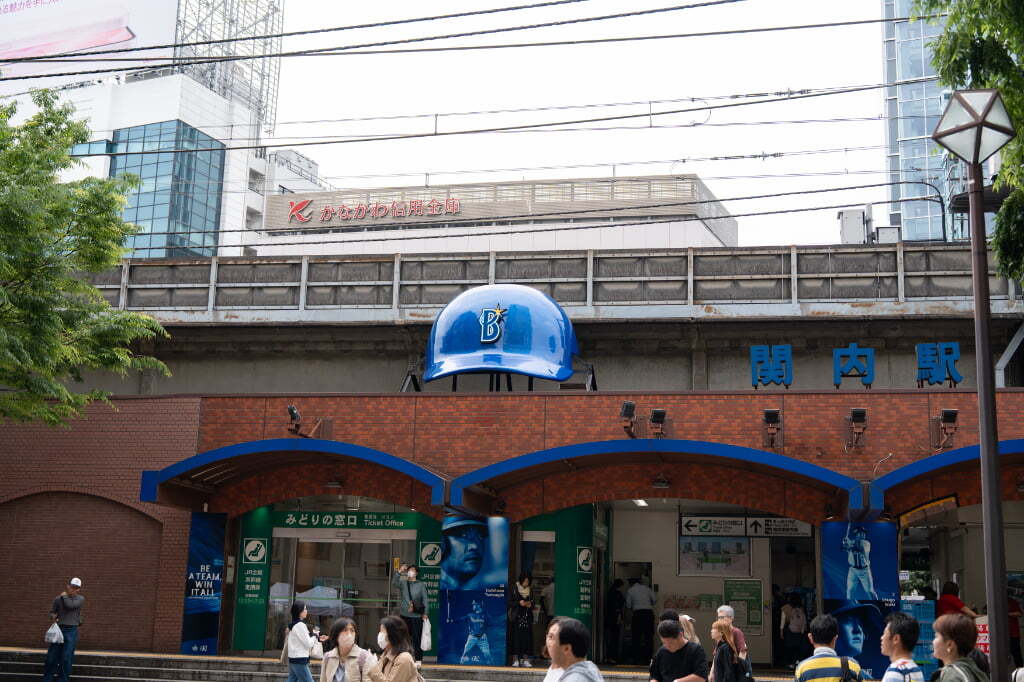 青いヘルメットの装飾が掲げられたJR関内駅の駅舎と、駅前を行き交う人