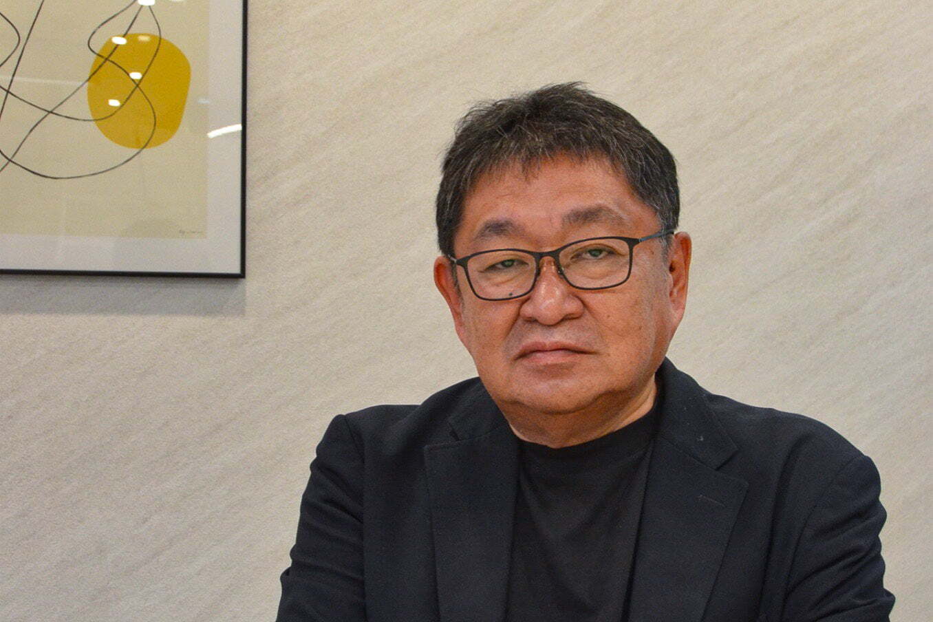 黄色い円と曲線の絵が掛かった壁の前で正面を向く、篠栗北地区産業団地の構想を民間側で率いた佐藤靖